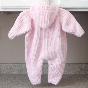 Puede incluir: Mono de beb&eacute; rosa claro con capucha, colgado de una percha blanca. El mono est&aacute; hecho de una tela de punto suave y texturizada. El mono con capucha tiene mangas largas y cubre las piernas.