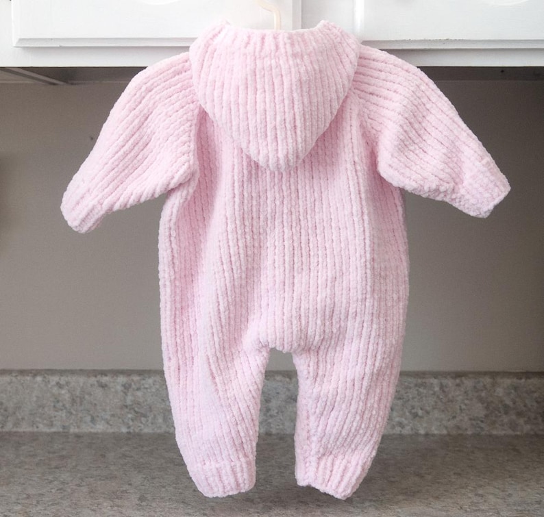 Puede incluir: Mono de beb&eacute; rosa claro con capucha, colgado de una percha blanca. El mono est&aacute; hecho de una tela de punto suave y texturizada. El mono con capucha tiene mangas largas y cubre las piernas.