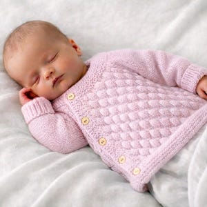 Könnte beinhalten: Ein schlafendes Baby trägt eine hellrosa Strickjacke mit einem strukturierten Muster und Holzknöpfen. Das Baby liegt auf einer weißen, strukturierten Decke. Die Strickjacke hat lange Ärmel und einen gerippten Kragen und Bündchen.
