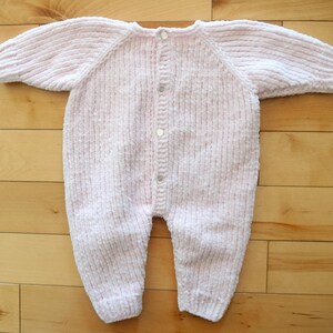 Puede incluir: Un pelele de beb&eacute; tejido en rosa claro, con mangas y piernas largas. El pelele tiene una botonadura con cuatro botones peque&ntilde;os y redondos. La tela parece suave y texturizada, adecuada para la delicada piel de un beb&eacute;.