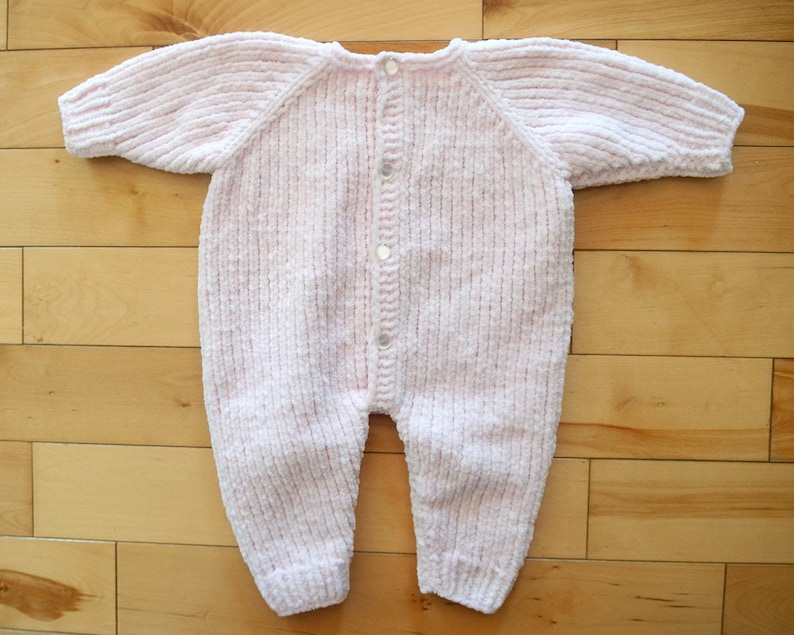 Puede incluir: Un pelele de beb&eacute; tejido en rosa claro, con mangas y piernas largas. El pelele tiene una botonadura con cuatro botones peque&ntilde;os y redondos. La tela parece suave y texturizada, adecuada para la delicada piel de un beb&eacute;.