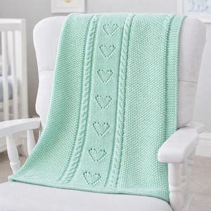 May include: A mint green knitted baby blanket with a central panel of heart-shaped cutouts and cable knit detailing. The blanket is draped over a white rocking chair in a nursery setting. The blanket is made of soft, textured yarn.