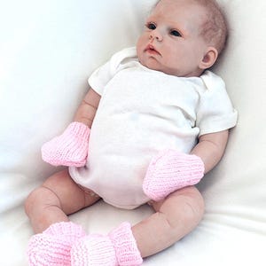 May include: A baby doll wearing a white onesie and pink knitted booties and mittens.