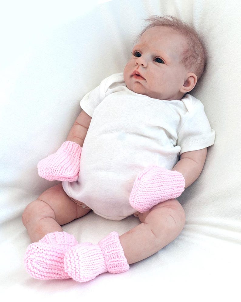 May include: A baby doll wearing a white onesie and pink knitted booties and mittens.