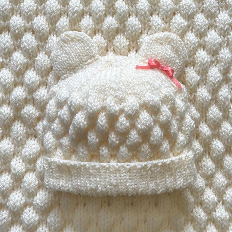 May include: Cream-colored knitted baby hat with bear ears and a pink bow. The hat features a textured, raised pattern and a folded brim. The hat is placed on a matching knitted blanket with a similar textured pattern.