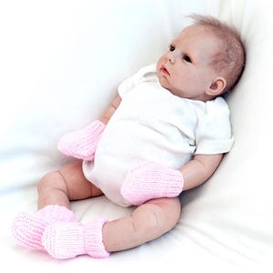 May include: A baby doll wearing a white onesie and pink knitted booties. The doll is lying on a white blanket.