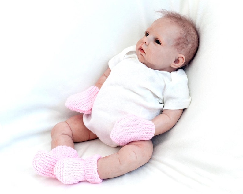 May include: A baby doll wearing a white onesie and pink knitted booties. The doll is lying on a white blanket.