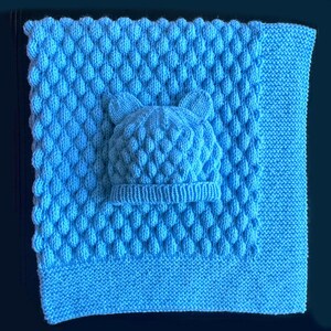 May include: A light blue knitted baby blanket and matching hat set. The blanket features a textured pattern, and the hat has bear ears. The set is made of soft yarn, perfect for a newborn.