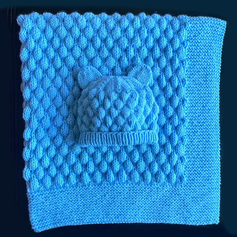 May include: A light blue knitted baby blanket and matching hat set. The blanket features a textured pattern, and the hat has bear ears. The set is made of soft yarn, perfect for a newborn.