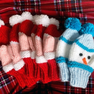 以下が含まれることがあります： 赤と白のニットミトン3組と、雪だるまのデザインの青と白のニットミトン1組のセットです。ピンクのアクセントが付けられています。