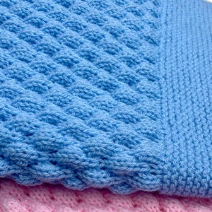May include: Close-up of a baby blue knitted blanket with a textured pattern. The blanket has a ribbed border and is layered over a pink knitted blanket. The soft, cozy texture is visible.