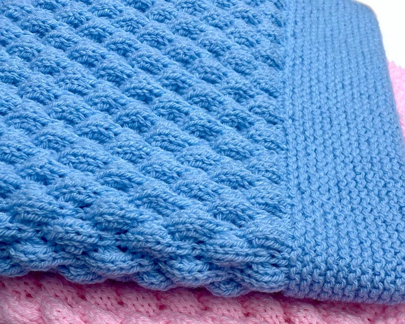 May include: Close-up of a baby blue knitted blanket with a textured pattern. The blanket has a ribbed border and is layered over a pink knitted blanket. The soft, cozy texture is visible.