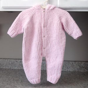 Puede incluir: Mono de beb&eacute; rosa claro con capucha, hecho de tejido de punto acanalado suave. La prenda de una pieza tiene mangas largas, cierre de botones en la parte delantera y pies adjuntos. Cuelga de una percha de pl&aacute;stico blanco.