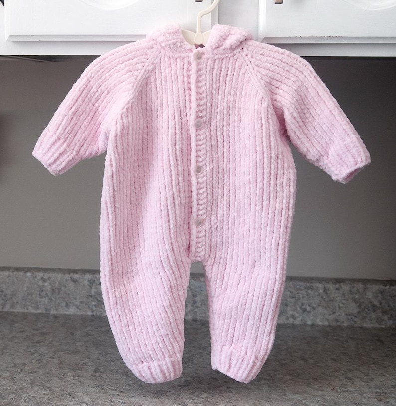 Puede incluir: Mono de beb&eacute; rosa claro con capucha, hecho de tejido de punto acanalado suave. La prenda de una pieza tiene mangas largas, cierre de botones en la parte delantera y pies adjuntos. Cuelga de una percha de pl&aacute;stico blanco.