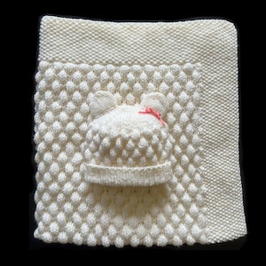 May include: Cream-colored knitted baby blanket and hat set. The blanket features a textured pattern, and the hat has bear ears and a small pink bow. The set is made of soft yarn, perfect for a newborn.