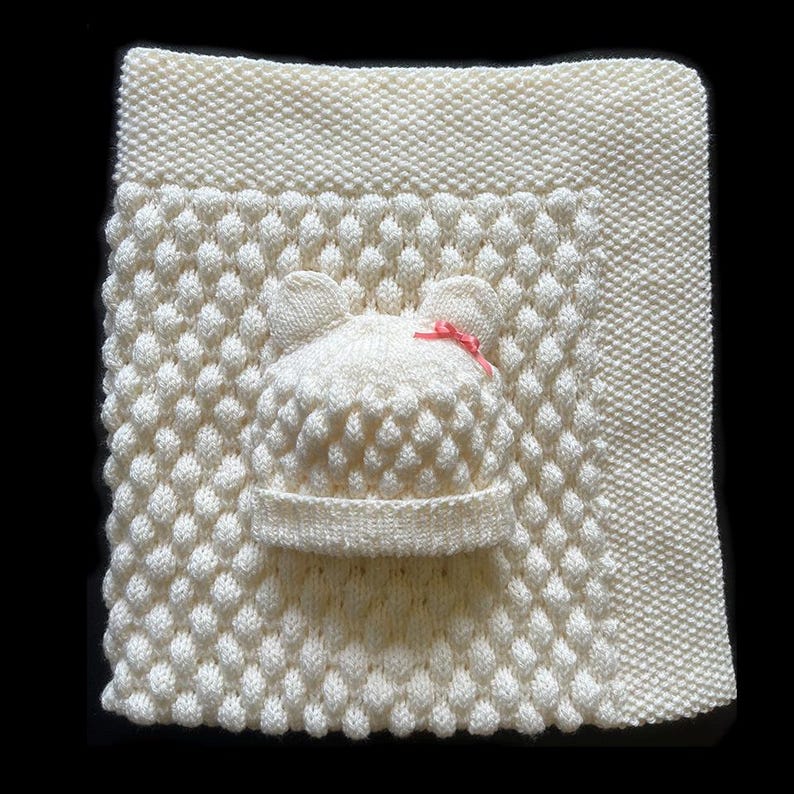 May include: Cream-colored knitted baby blanket and hat set. The blanket features a textured pattern, and the hat has bear ears and a small pink bow. The set is made of soft yarn, perfect for a newborn.