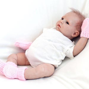 May include: A pink baby doll wearing a white onesie and pink knitted booties and mittens.