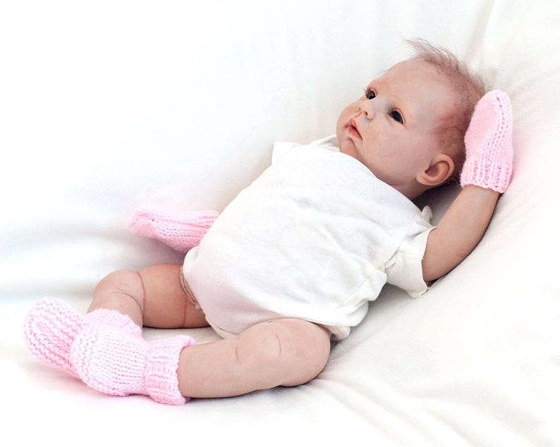 May include: A pink baby doll wearing a white onesie and pink knitted booties and mittens.