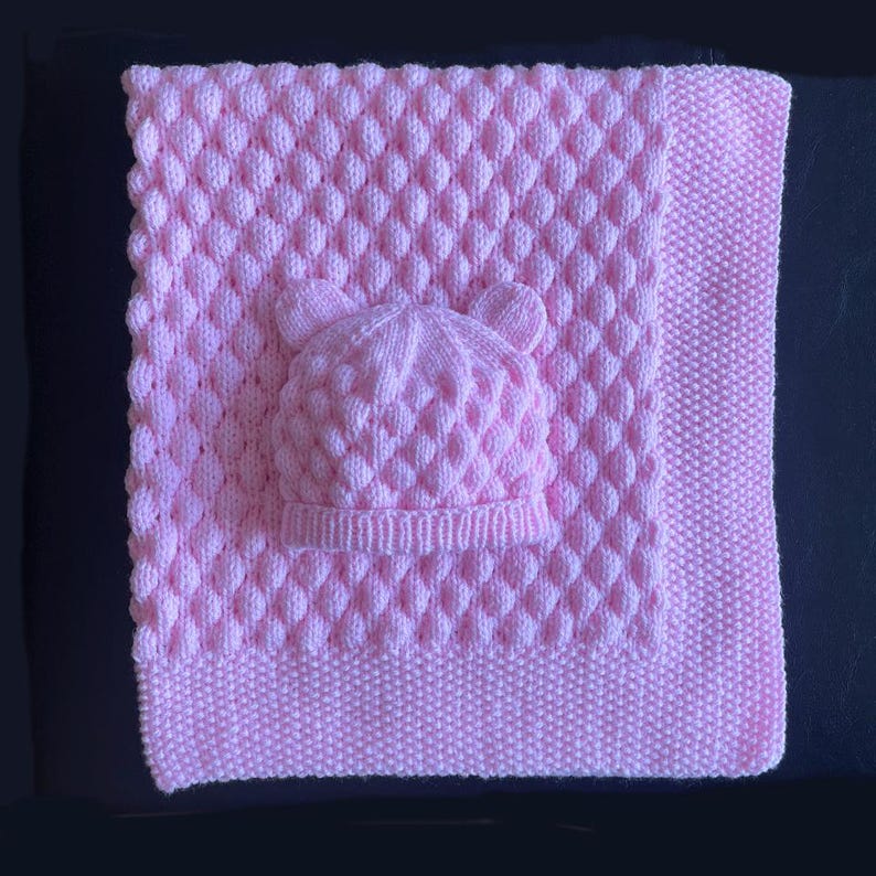 May include: A soft pink knitted baby blanket and matching hat set. The blanket features a textured, diamond-like pattern, while the hat has small bear ears. The set is made of a soft, possibly wool, material.