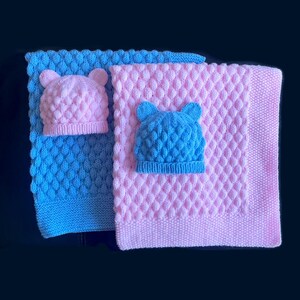 May include: Two knitted baby blankets, one blue and one pink, each with a matching hat. The hats have bear ears. The blankets have a textured diamond pattern. The items are likely handmade.