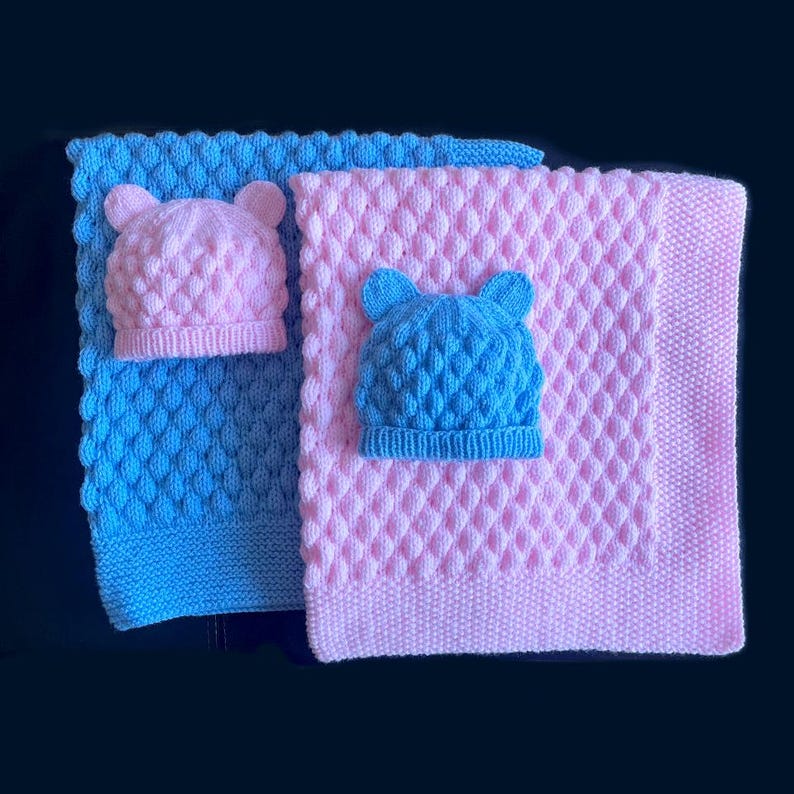 May include: Two knitted baby blankets, one blue and one pink, each with a matching hat. The hats have bear ears. The blankets have a textured diamond pattern. The items are likely handmade.