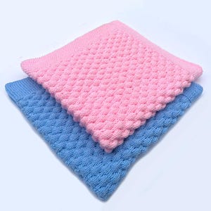May include: Two knitted baby blankets, one pink and one blue, are stacked diagonally. The blankets have a textured, raised pattern and a scalloped edge. The pink blanket is on top, partially covering the blue blanket.