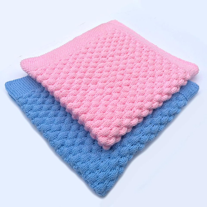 May include: Two knitted baby blankets, one pink and one blue, are stacked diagonally. The blankets have a textured, raised pattern and a scalloped edge. The pink blanket is on top, partially covering the blue blanket.