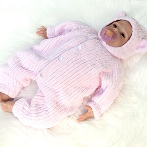Puede incluir: Una mu&ntilde;eca beb&eacute; realista vestida con un traje de una pieza rosa claro texturizado y una capucha a juego con orejas peque&ntilde;as. La mu&ntilde;eca tiene un chupete en la boca y est&aacute; acostada sobre una superficie blanca y esponjosa.