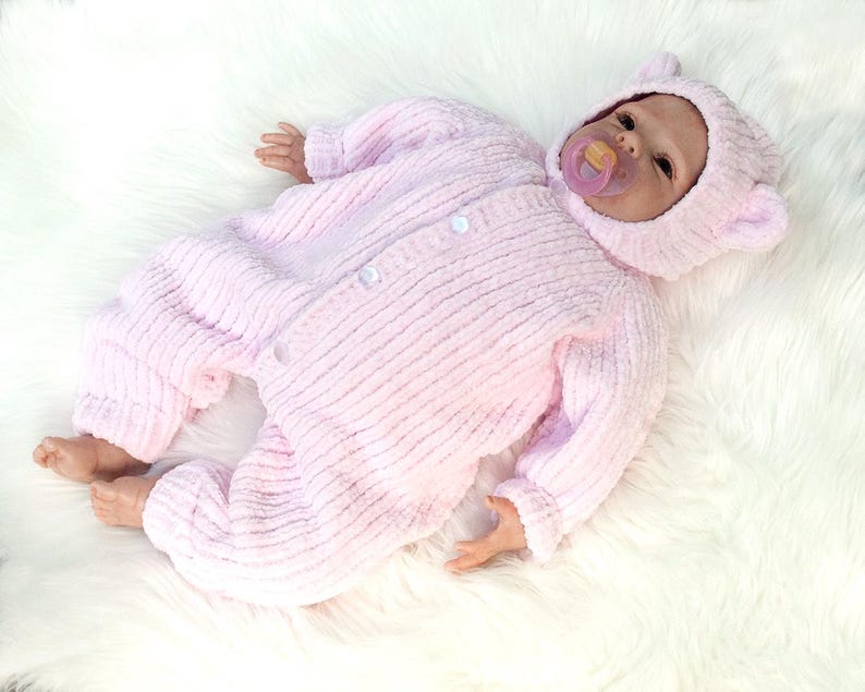 Puede incluir: Una mu&ntilde;eca beb&eacute; realista vestida con un traje de una pieza rosa claro texturizado y una capucha a juego con orejas peque&ntilde;as. La mu&ntilde;eca tiene un chupete en la boca y est&aacute; acostada sobre una superficie blanca y esponjosa.
