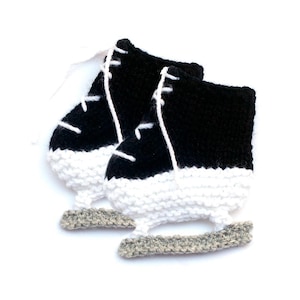May include: A pair of black and white knitted ice skate booties. The booties are made with a white yarn and have black knit details. The booties have a gray knit detail that resembles the blade of an ice skate.
