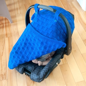 Op de afbeelding: Een blauwe gebreide deken bedekt een babyautostoeltje met een baby erin. De autostoel is zwart met een grijs-wit gestreept patroon.