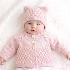 Op de afbeelding: Een baby draagt een lichtroze gebreid vest en een bijpassende muts met oor details. Het vest heeft een textuur van een ruitpatroon op de onderkant van de voorkant en is dichtgeknoopt. De baby ligt op een witte ondergrond.