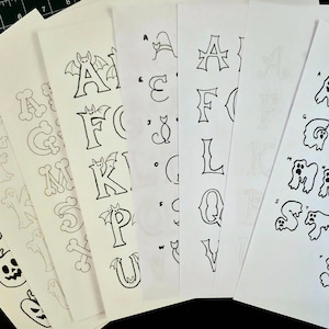 Puede incluir: Una colección de siete hojas de papel blanco con ilustraciones en tinta negra de letras del alfabeto. Los diseños incluyen letras con forma de fantasma, tipografía de huesos, caracteres adornados con murciélagos y motivos de Halloween.