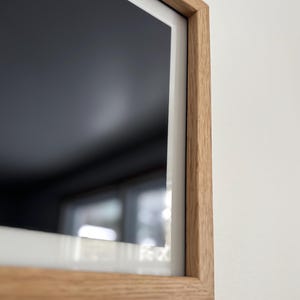 May include: A rectangular, modern television with a light oak wood frame. The screen is black, and the frame has a white border. The TV is mounted on a white wall.