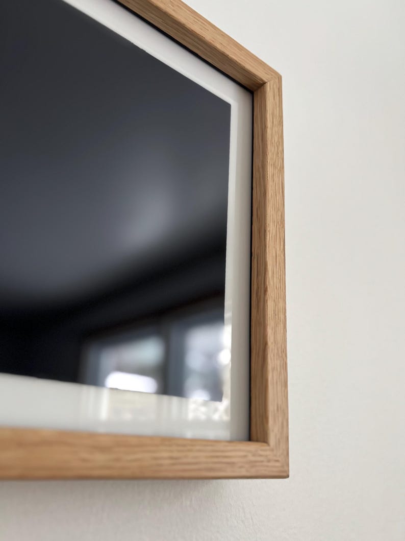 May include: A rectangular, modern television with a light oak wood frame. The screen is black, and the frame has a white border. The TV is mounted on a white wall.