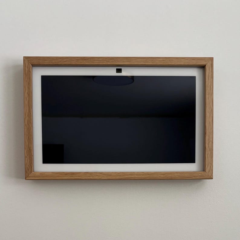 May include: A rectangular digital photo frame with a light oak wood frame. The screen is black, with a white border. A small, dark circular camera is centered at the top. The frame is mounted on a white wall.