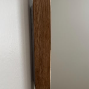 May include: A side view of a rectangular wooden frame with a natural wood grain finish. The frame is mounted on a white wall. The wood has a warm, brown tone.