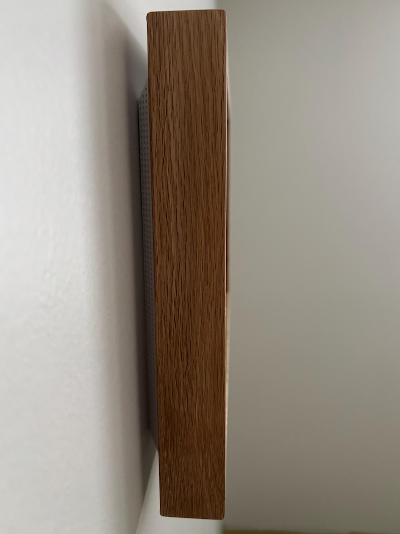 May include: A side view of a rectangular wooden frame with a natural wood grain finish. The frame is mounted on a white wall. The wood has a warm, brown tone.