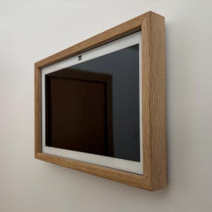 May include: A rectangular digital display with a black screen is mounted on a white wall. The display is encased in a light brown wooden frame. The frame has a natural wood grain texture, adding a touch of warmth to the modern design.