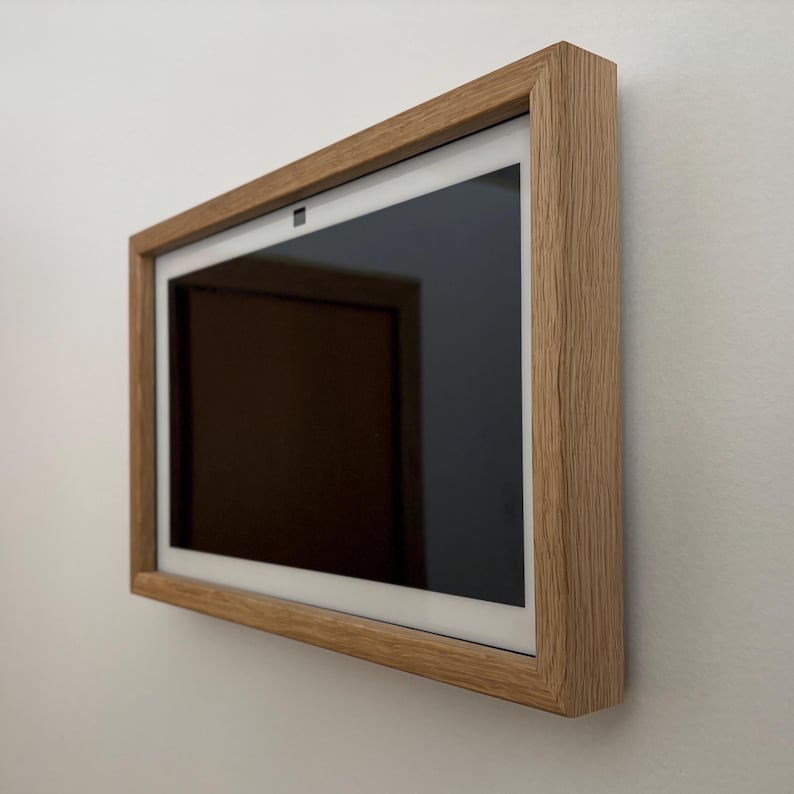 May include: A rectangular digital display with a black screen is mounted on a white wall. The display is encased in a light brown wooden frame. The frame has a natural wood grain texture, adding a touch of warmth to the modern design.