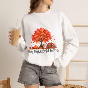 May include: A white long-sleeve sweatshirt featuring a graphic of an autumn tree with orange and red leaves, a pumpkin, a basket of apples, and a mug. The text below the illustration reads Cozy Days, Colorful Leaves.