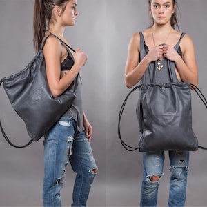 May include: Two models are wearing a grey leather drawstring backpack. The backpack has a long strap and can be worn as a shoulder bag or a backpack. The backpack is made of soft, supple leather and has a simple, elegant design.