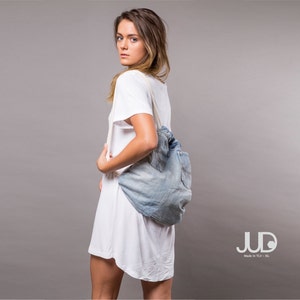 May include: A woman wearing a white t-shirt dress with a denim tote bag slung over her shoulder. The bag has a white strap and is made of recycled denim. The bag is a light blue color and has a slightly distressed look. The woman is looking over her shoulder at the camera.
