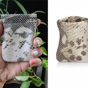 May include: A small, beige and brown snakeskin pouch with a drawstring closure. The pouch is made of a soft, supple leather and has a subtle sheen.