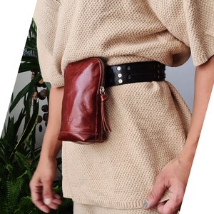 May include: A brown leather fanny pack with a zipper closure and a black studded belt. The fanny pack is worn over a beige sweater.