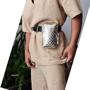 K&ouml;nnte beinhalten: Eine silberne und goldene G&uuml;rteltasche mit wei&szlig;en Punkten und einem schwarzen Gurt. Die G&uuml;rteltasche wird von einer Person getragen, die einen beigefarbenen Pullover tr&auml;gt.