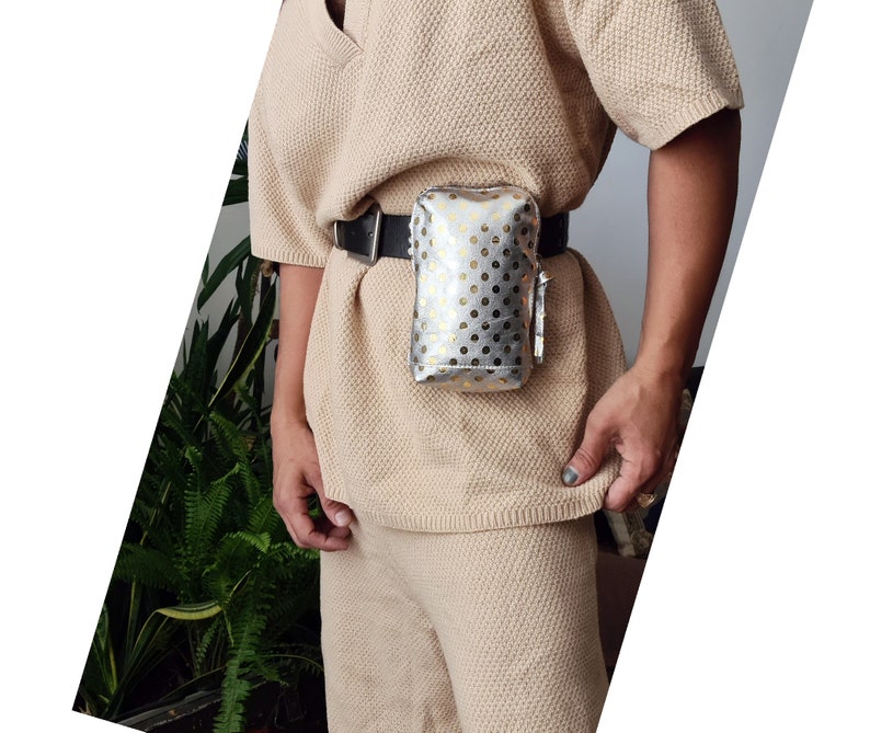 K&ouml;nnte beinhalten: Eine silberne und goldene G&uuml;rteltasche mit wei&szlig;en Punkten und einem schwarzen Gurt. Die G&uuml;rteltasche wird von einer Person getragen, die einen beigefarbenen Pullover tr&auml;gt.
