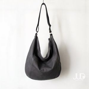 May include: A large, dark grey leather tote bag with a long shoulder strap. The bag has a slouchy, casual look and is hanging from a hook.