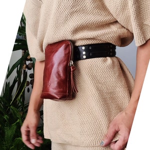 K&ouml;nnte beinhalten: Eine braune Leder-Bauchtasche mit Rei&szlig;verschluss und einem schwarzen G&uuml;rtel mit Nieten. Die Bauchtasche wird &uuml;ber einem beigefarbenen Pullover getragen.