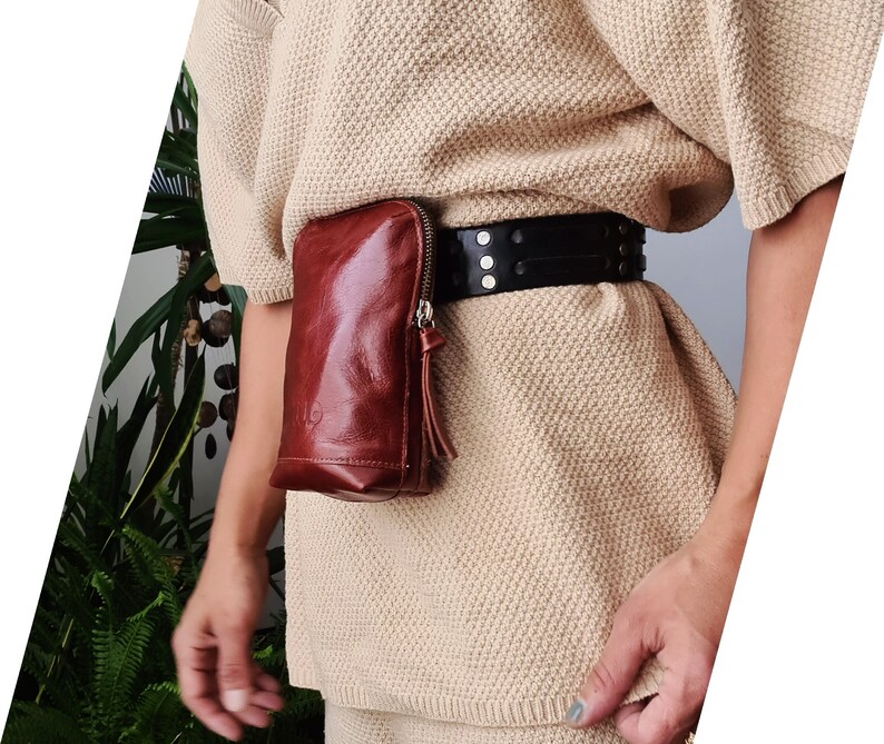 K&ouml;nnte beinhalten: Eine braune Leder-Bauchtasche mit Rei&szlig;verschluss und einem schwarzen G&uuml;rtel mit Nieten. Die Bauchtasche wird &uuml;ber einem beigefarbenen Pullover getragen.
